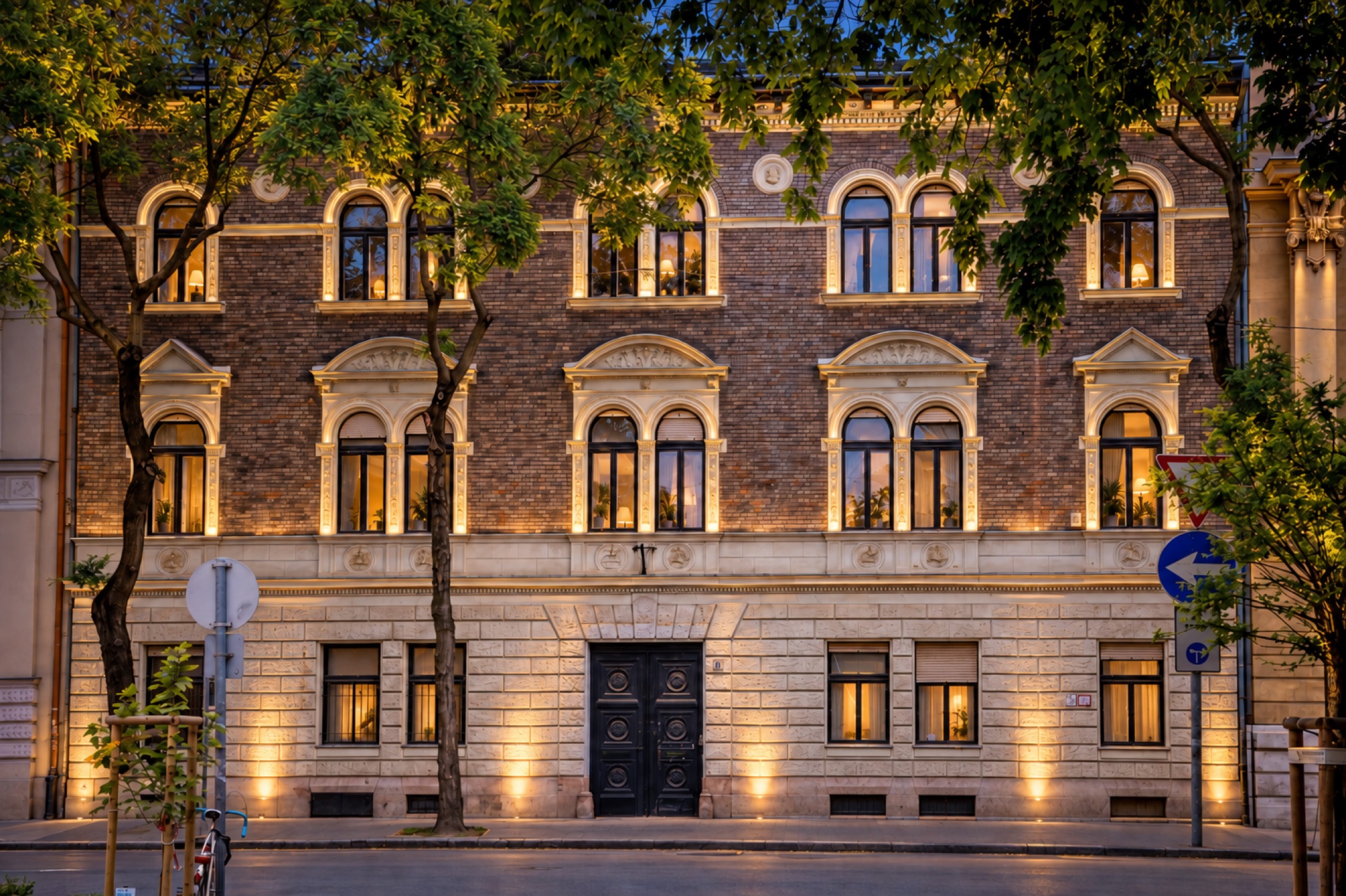The restored street façade at dusk: brick above a rusticated stone base, arched windows with carved surrounds, lit from below, framed by street trees
